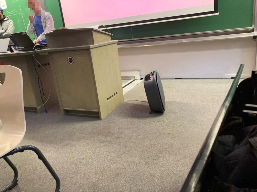 A photo taken at FOSDEM showing an air filter device on the floor near a speaker in the Policy Dev Room