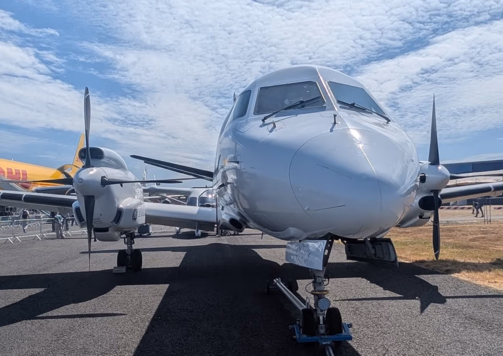 GE Aerospace's hybrid-electric Saab 340B testbed aircraft on static display at an airshow, viewed head-on. The asymmetric engine configuration is clearly visible: on the left wing, the hybrid-electric CT7 turboprop nacelle carries a smaller GE-branded electric motor pod mounted beneath it, while the right wing carries the standard unmodified CT7 turboprop. Both engines drive Dowty multi-blade propellers. A DHL cargo aircraft is visible in the background to the left